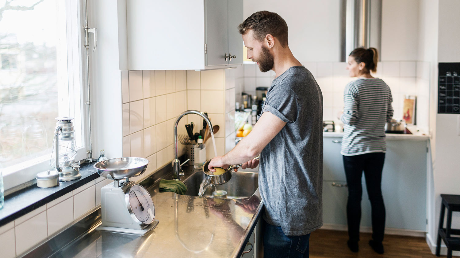 Man washing pan while wife stands in background of kitchen